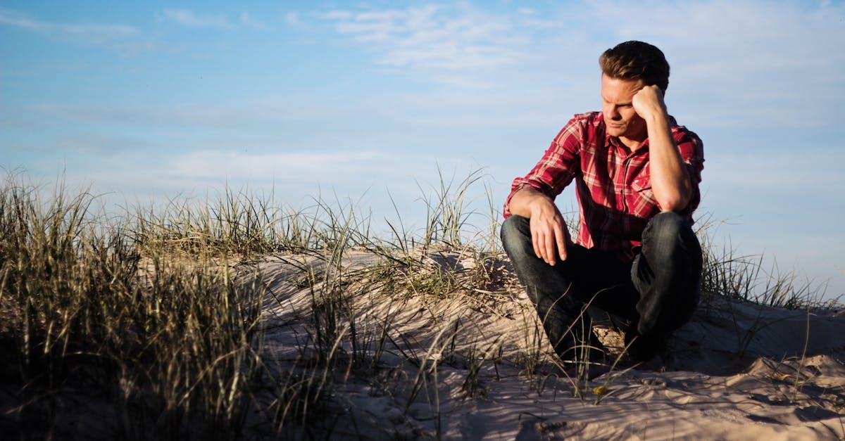 Man in a red plaid shirt sitting thoughtfully on a sandy dune in Australia, surrounded by grass.