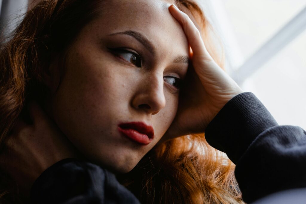 Žena zamišljeno gleda kroz prozor dok rukom pridržava glavu, prikazujući potisnutu grižnju savesti, unutrašnji nemir i emotivnu težinu.