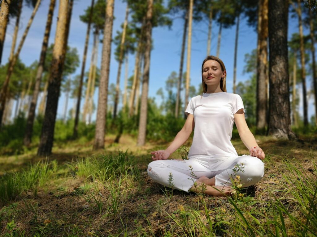 A woman in white sits in lotus pose, meditating in a serene forest setting.
