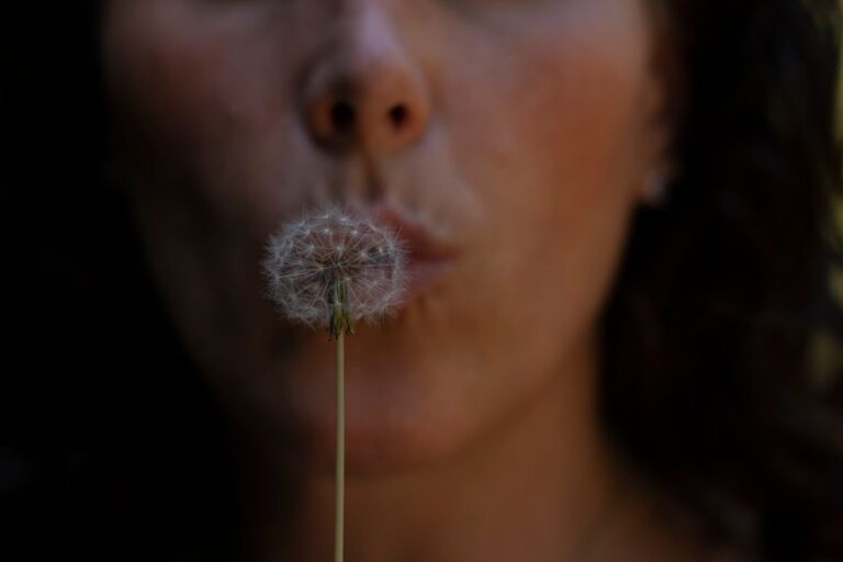 A close-up view of a woman blowing dandelion seeds outdoors in soft focus.